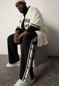 Beige Baseball-Trikot mit schwarzen Akzenten, kombiniert mit schwarzen Hosen mit weißen Druckknöpfen und Streifen, getragen mit weißen Turnschuhen.