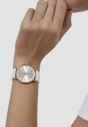 Runde Uhr mit weißem Silikonarmband und silbernem Zifferblatt. Verfügt über roségoldene Stundenmarkierungen und eine gepunktete Lünette. Einfaches und modernes Design.