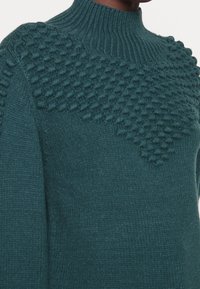 Pull turquoise à col montant, motif en relief à points boules sur la partie supérieure, et tricot lisse sur les manches.