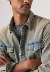 Άνδρας που φοράει ξεθωριασμένο τζιν μπουφάν πάνω από γκρι πουκάμισο με ασημένιο κολιέ αλυσίδα, με σταυρωμένα τα χέρια, κοιτάζοντας προς τα πλευρά μπροστά σε απλό φόντο.