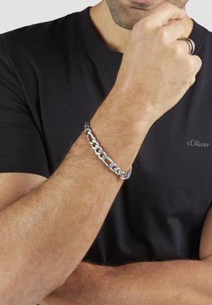 Mann trägt ein schwarzes s.Oliver T-Shirt mit einem silbernen Gliederarmband und einem schwarzen und silbernen Ring an der Hand in der Nähe seines Gesichts.