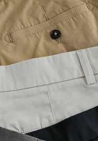 Pantalons empilés beige, gris clair et noir montrant les détails des coutures, un bouton, une boucle de ceinture et des ouvertures de poches.