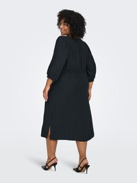 Musta polvipituinen mekko, jossa on smokkivyötärö, lyhyet puffihihaiset hihat ja sivuhalkiot. Malli käyttää mustia remmikorkokenkiä. Kankaan pinta on teksturoitu.