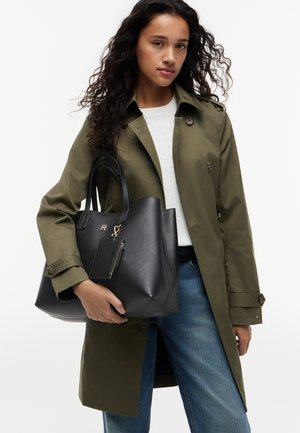 Mujer con gabardina de color oliva y suéter blanco, sosteniendo un gran bolso tote negro de cuero con un pequeño monedero adjunto.