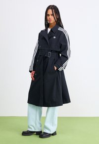 Trench-coat noir à ceinture avec poches latérales, présentant des accents blancs à trois bandes sur les manches ; associé à un pantalon large bleu clair et des talons épais noirs.