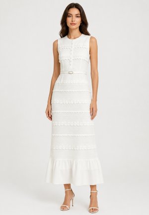 Vrouw draagt een witte mouwloze midi-jurk met bloemmotieven en geschulpte details, een riem, en witte sandalen met hoge hakken op een effen achtergrond.