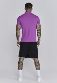 Paarse aangesloten t-shirt met korte mouwen en zwarte shorts, met tatoeages zichtbaar op de armen en benen, gecombineerd met witte sneakers en crew sokken.