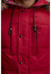 Veste matelassée rouge avec capuche bordée de fourrure, dotée de cordons noirs, de boutons-pression et de deux poches avant avec motifs de couture horizontaux.