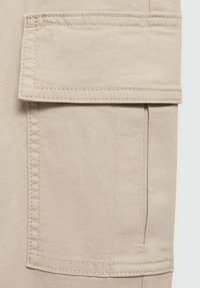 Beige tygficka med klaff på ett par byxor, med synlig sömnad och en slät yta. Enkel design utan mönster.