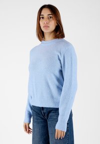Maglione azzurro chiaro, di texture, con scollo rotondo e polsini a costine, abbinato a jeans di denim scuro. Il tessuto appare morbido e accogliente.