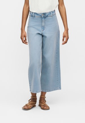 Person iført lys blå vidde korte jeans, hvid top og brune flade sandaler med remme, stående mod en ensfarvet baggrund.