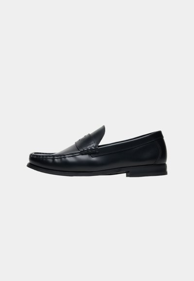 Μαύρο δερμάτινο penny loafer με χαμηλό τακούνι και λεπτομερή ραφή κατά μήκος της άνω άκρης, εμφανιζόμενο από το πλάι σε απλό φόντο.