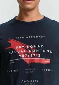 Camiseta de algodón azul marino con un diseño gráfico rojo de una aeronave y texto blanco que detalla temas de aviación, incluyendo "Team Aeronaut."
