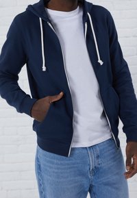 Marinblå zip-up hoodie med vita snören, kängurufickor och ribbad mudd, bärs över en vit t-shirt och kombineras med ljusblå jeans.