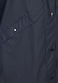 Section de tissu d'une veste bleu marine foncé montrant une poche à rabat inclinée avec bouton-pression et une ligne verticale de trois boutons-pression noirs.