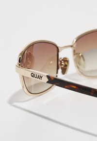 Gafas de sol en tono dorado con lentes marrones de degradado, que cuentan con una patilla con patrón de carey y el logo grabado "QUAY" en la varilla.