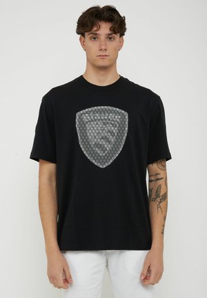 Giovane uomo con capelli ricci, che indossa una maglietta nera con emblema a scudo bianco a motivo a diamante, pantaloni bianchi e tatuaggi visibili sulle braccia, in piedi su sfondo bianco.