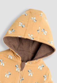 Giacca gialla con cappuccio e rivestimento a coste marrone, decorata con piccole illustrazioni di zebra bianche e nere, cerniera frontale visibile.