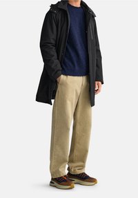 Sort vandtæt frakke, mørkeblå strikket sweater, beige bukser med brede ben og multifarvede sneakers med orange detaljer. Afslappet, lagdelt outfit.