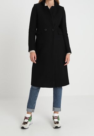 Femme portant un manteau noir mi-long par-dessus un jean bleu roulé et des baskets blanches avec des accents verts, debout devant un fond uni.
