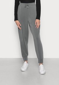 Person klädd i grå joggingbyxor med dragsko och vita sneakers, stående mot en enkel ljus bakgrund.