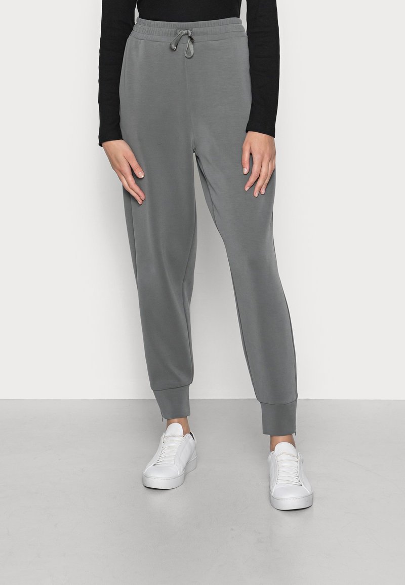 Person klädd i grå joggingbyxor med dragsko och vita sneakers, stående mot en enkel ljus bakgrund.
