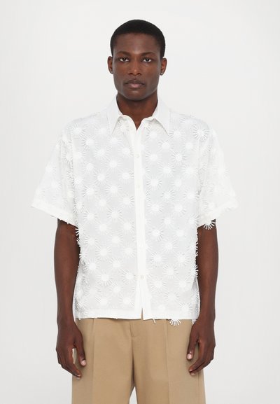 Chemise blanche à manches courtes avec des motifs de soleil brodés, patte de boutonnage sur le devant et texture douce. Associée à un pantalon beige.