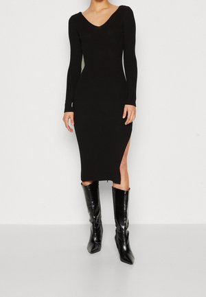 Kobieta ubrana w dopasowaną czarną sukienkę midi z długimi rękawami i bocznym rozcięciem, w parze z błyszczącymi czarnymi, sięgającymi kolan butami na szpic.