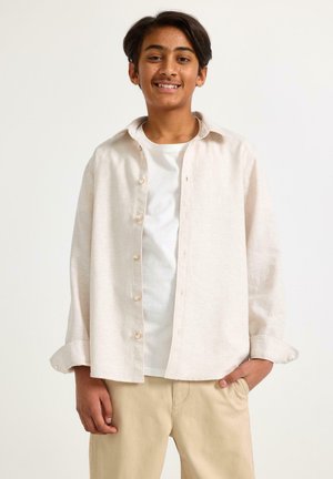 Teini-ikäinen poika hymyilemässä, päällään avoin kermaan vivahtava napitettava paita valkoisen T-paidan päällä ja beiget housut, seisomassa yksinkertaisen taustan edessä.