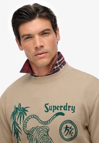 Uomo che indossa un felpa beige di Superdry con grafiche di tigre verde e colletto di camicia a quadri rossi visibile, sfondo neutro.