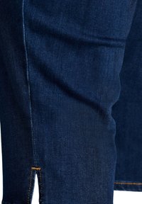 Σκούρα μπλε τζιν από denim που δείχνει κοντινή λήψη μιας πλευρικής ραφής με πορτοκαλί ραφή και μικρή κάτω σχισμή.