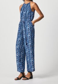 Blå mönstrad jumpsuit med halterneck, insvängd midja och vida ben. Har blommönster och tofsar. Matchad med svarta sandaletter med remmar.