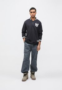 Tengerészkék pulóver fehér logóval, csíkos gallérral. Szürke bőszárú nadrág, bézs csizma. Egyszerű kialakítás, kényelmes szabás, sima anyag és minimális részletek.