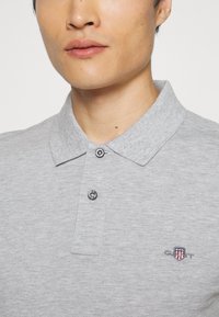 Grå polo skjorte lavet af blødt stof med en struktureret krave, to knaplukninger og broderet logo med et skjolddesign.