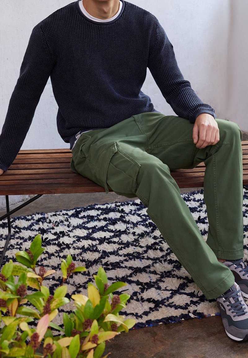 Gröna cargobyxor i kombination med en svart strukturerad stickad tröja. Modellen sitter på en bänk i trä, med grå sneakers och en mönstrad matta nedanför.