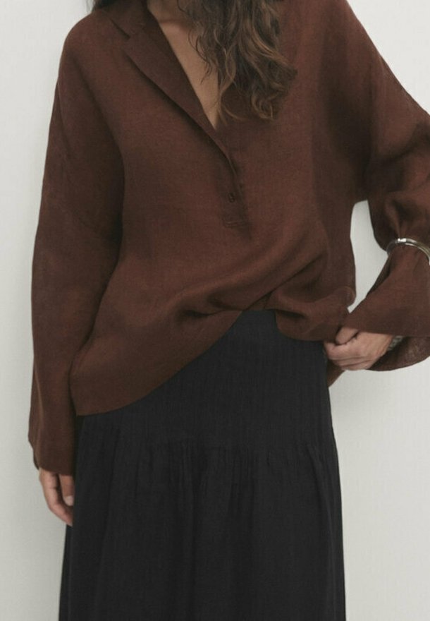 Femme portant un chemisier marron ample à col en V et manches longues, rentré dans une jupe noire fluide taille haute, avec un bracelet argenté au poignet.