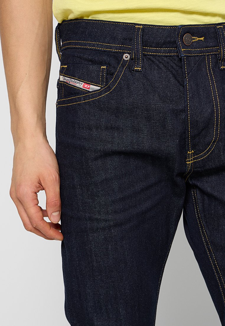 Dunkelblaue Jeans mit gelben Nähten, ausgestattet mit einer Vordertasche mit einem gebrandeten Etikett und einem Knopfverschluss an der Taille.
