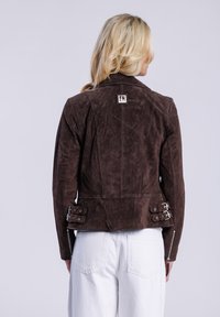 Giacca in suede marrone con design aderente, caratterizzata da accessori in metallo argentato, un'etichetta sul retro e cinturini laterali regolabili.