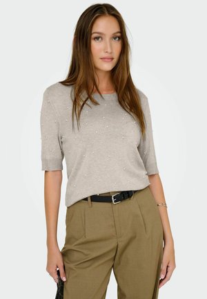 Femme avec de longs cheveux bruns portant un pull beige à manches courtes orné de petites perles et un pantalon olive taille haute avec une ceinture noire.