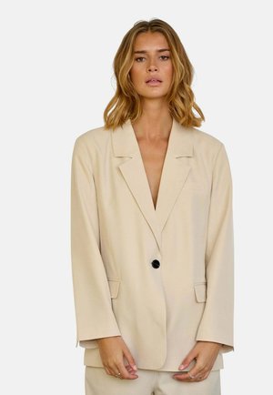 Bézs blézer hajtókkal, egy fekete gombbal záródik, és oldalzsebekkel rendelkezik. Simaságú anyagból készült, szabott fazonban és laza ujjak.