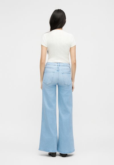 Γυναίκα με μακριά σκούρα μαλλιά που φοράει λευκό κοντομάνικο τοπ και ανοιχτό μπλε τζιν παντελόνι με φαρδιά μπατζάκια, όρθια με την πλάτη προς το μέρος μας σε απλό φόντο.