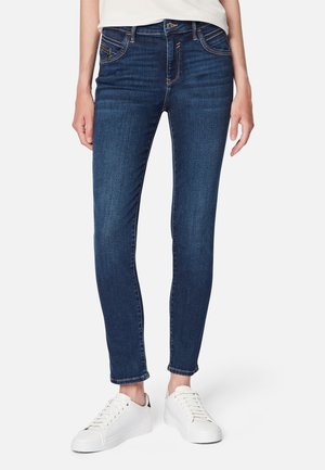 Jean slim fit en denim bleu foncé porté avec des baskets blanches sur une personne debout devant un fond uni.