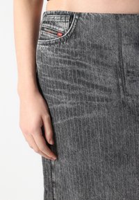Grå denimskjørt med vertikale stripes, en frontlomme med logomerking, og en lett teksturert overflate. Hånden hviler på stoffet.