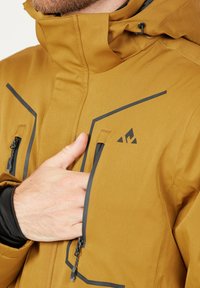 Brun vattentät jacka med hög krage, grå accentdetaljer, dragkedjefickor och en logotyp på bröstet. Hållbar tygtextur.