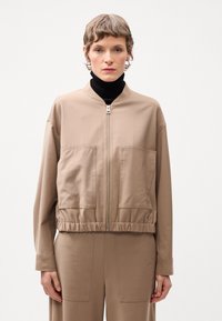 Beige bomberjacka med ribbad nederkant, dragkedja och två fickor fram. Slät tyg, avslappnad passform och hög kragodesign.