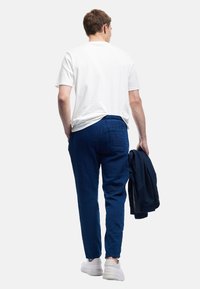 Weißes Kurzarm-T-Shirt, marineblaue strukturiertes Hose mit elastischem Bund, getragen mit weißen Sneakers, hält eine dunkle Jacke.