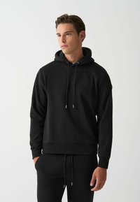 Sudadera negra hecha de una tela suave y texturizada. Presenta una capucha con cordón, puños acanalados y una cinturilla ajustada para un aspecto aerodinámico.