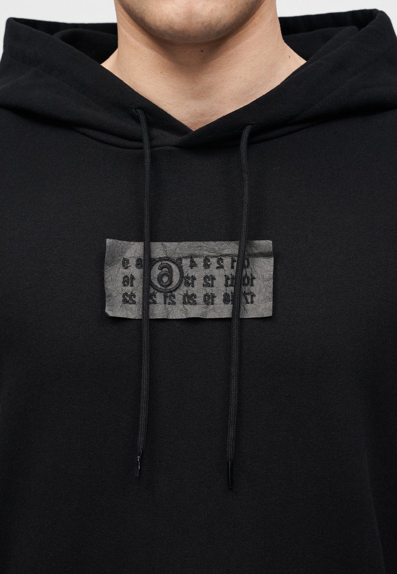Mann trägt schwarzen Hoodie mit schwarzen Kordeln und einem strukturierten grauen Aufnäher mit abstraktem schwarzen Text auf der Brust.