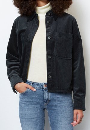 Femme portant une veste en velours côtelé noire à boutons par-dessus un pull à col roulé blanc et un jean denim bleu clair, avec une main dans la poche.