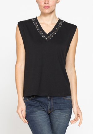 Donna che indossa un top nero senza maniche con scollo a V decorato con perline e jeans blu, in piedi davanti a uno sfondo bianco uniforme.